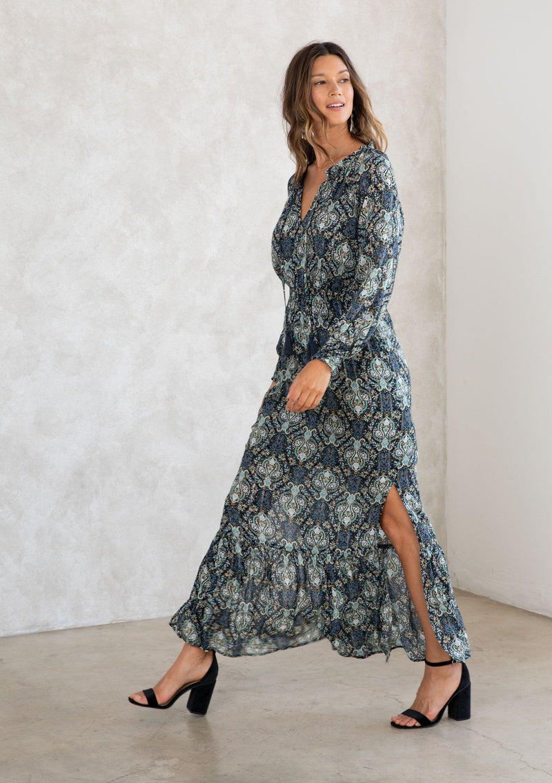 [Color: Black/Teal] A model wearing a black and teal paisley print maxi dress with light catching lurex thread. With long voluminous sleeves, a split neckline with tassel ties, side slits, and an elastic waist. 