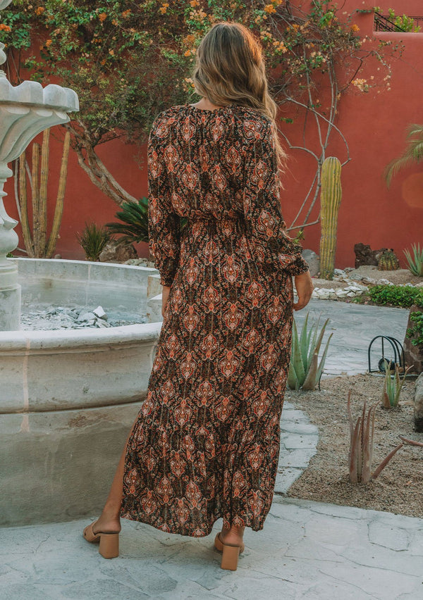 [Color: Black/Cayenne] A model wearing a black and red paisley print maxi dress with light catching lurex thread. With long voluminous sleeves, a split neckline with tassel ties, side slits, and an elastic waist. 