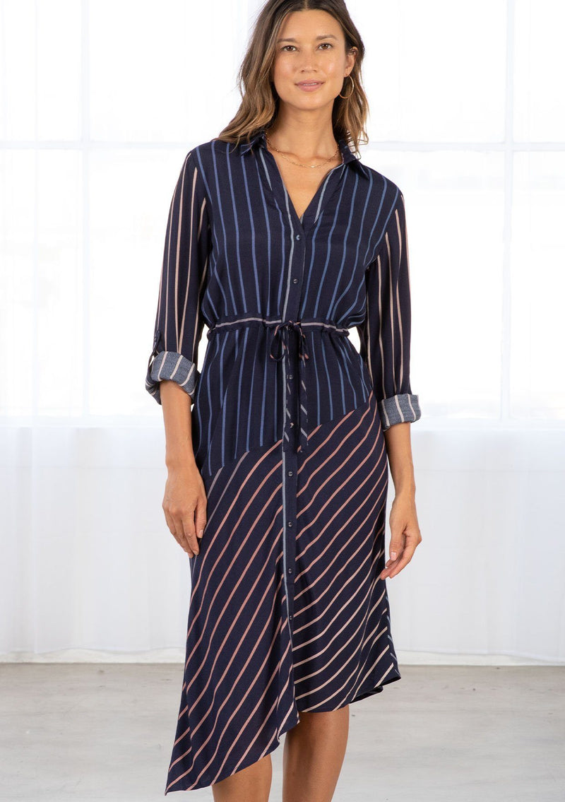 [Color: Navy/Blue] A model wearing a navy blue shirt dress in a gradient stripe print. With long rolled sleeves and a button tab closure, an adjustable drawstring waist, a classic collared neckline, a button front, and an asymmetric mid length hemline. 