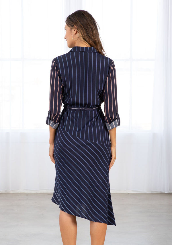 [Color: Navy/Blue] A model wearing a navy blue shirt dress in a gradient stripe print. With long rolled sleeves and a button tab closure, an adjustable drawstring waist, a classic collared neckline, a button front, and an asymmetric mid length hemline. 