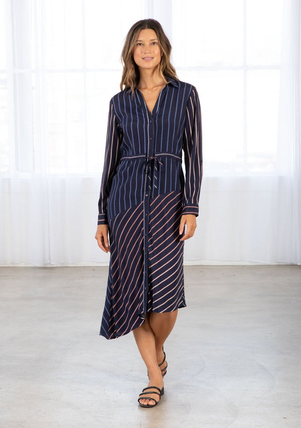 [Color: Navy/Blue] A model wearing a navy blue shirt dress in a gradient stripe print. With long rolled sleeves and a button tab closure, an adjustable drawstring waist, a classic collared neckline, a button front, and an asymmetric mid length hemline. 