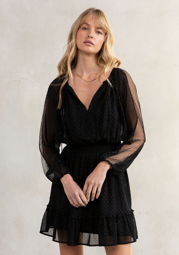 [Color: Black] A model wearing a black bohemian mini dress in a dot chiffon. With embroidered mesh trimmed long sleeves, an embroidered front detail, a smocked elastic waist, a split v neckline with ties, and a ruffle trimmed tiered skirt.