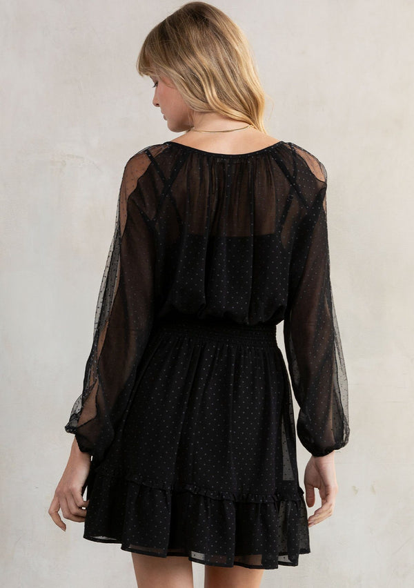 [Color: Black] A model wearing a black bohemian mini dress in a dot chiffon. With embroidered mesh trimmed long sleeves, an embroidered front detail, a smocked elastic waist, a split v neckline with ties, and a ruffle trimmed tiered skirt.