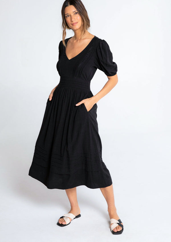 [Color: Black] A model wearing a timeless black linen blend maxi dress. With short puff sleeves, a flattering pleated waist, and delicate lattice trim.