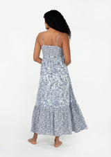 [Color: Ivory/Blue] A back facing image of a brunette model wearing a bohemian spring resort maxi dress in an ivory and blue mixed floral and paisley print. With adjustable spaghetti straps, a straight neckline, a slim fit smocked bodice, a button front, a flowy tiered skirt, and side pockets. 