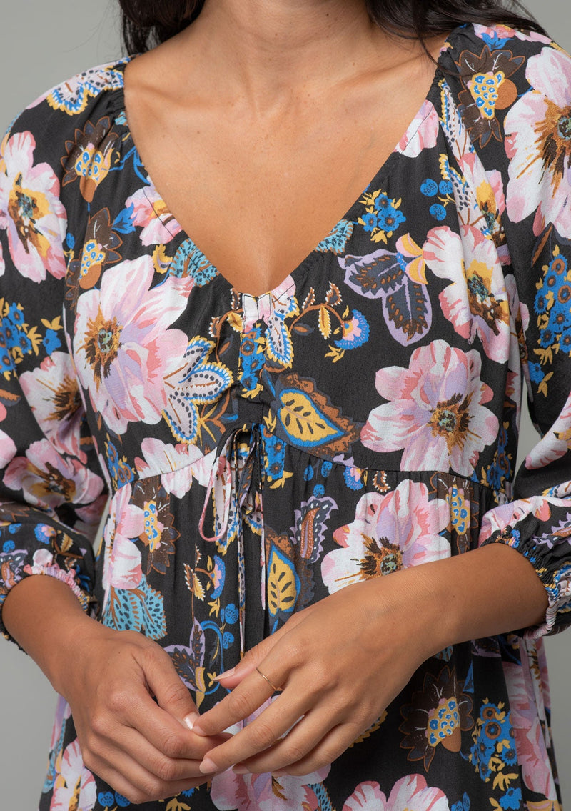 [Color: Charcoal/Rose] A close up front facing image of a brunette model wearing a charcoal grey and pink floral dress with a drawstring v neckline, a tiered high low hemline, and a flowy baby doll silhouette. 