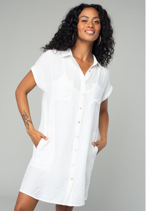 [Color: White] A front facing image of a brunette model wearing a loose and relaxed fit white mini shirt dress. With short cuffed sleeves, a button front, front patch pockets, and side pockets. 