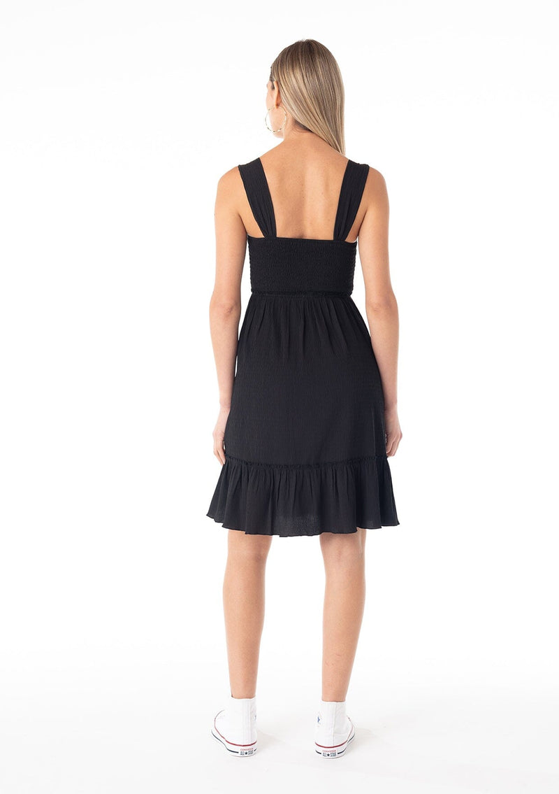 [Color: Black] A back facing image of a blonde model wearing a black sleeveless spring mini dress. With wide tank top straps, a square neckline, a smocked slim fit bodice, an empire waist, and a ruffle trimmed tiered skirt.