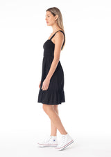 [Color: Black] A side facing image of a blonde model wearing a black sleeveless spring mini dress. With wide tank top straps, a square neckline, a smocked slim fit bodice, an empire waist, and a ruffle trimmed tiered skirt.