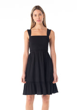 [Color: Black] A front facing image of a blonde model wearing a black sleeveless spring mini dress. With wide tank top straps, a square neckline, a smocked slim fit bodice, an empire waist, and a ruffle trimmed tiered skirt.