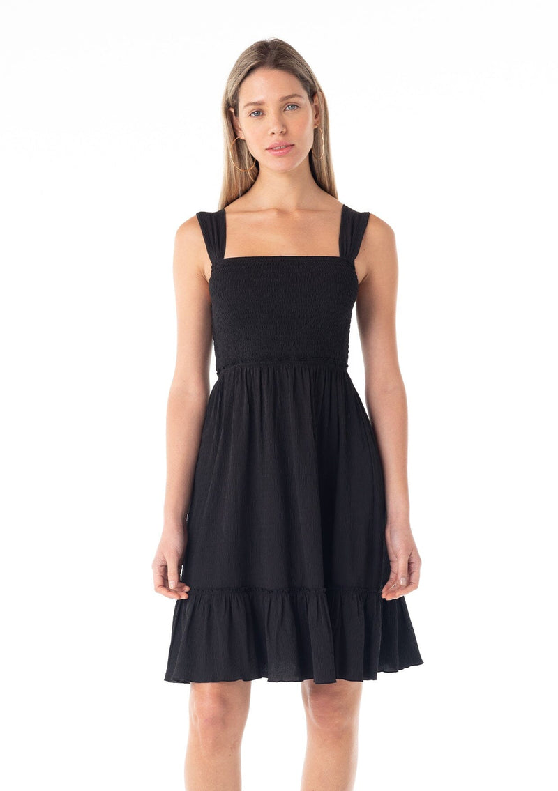 [Color: Black] A front facing image of a blonde model wearing a black sleeveless spring mini dress. With wide tank top straps, a square neckline, a smocked slim fit bodice, an empire waist, and a ruffle trimmed tiered skirt.