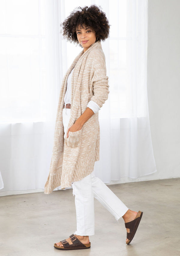 [Color: Nude/Ivory] A model wearing a beige knit reversible cardigan. With long sleeves, a waterfall open front, side pockets, and a shawl collar. 