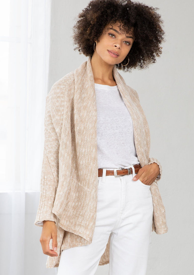 [Color: Nude/Ivory] A model wearing a beige knit reversible cardigan. With long sleeves, a waterfall open front, side pockets, and a shawl collar. 