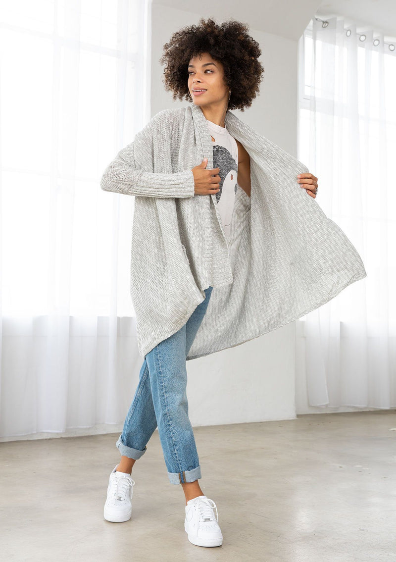 [Color: Silver/Ivory] A model wearing a silver grey knit reversible cardigan. With long sleeves, a waterfall open front, side pockets, and a shawl collar. 