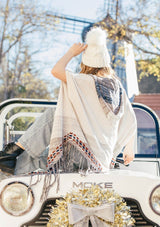 [Color: Sand/Grey] A blond woman wearing a lightweight hooded poncho. Featuring a fringed asymmetric hemline, an oversize hood, and a multi color striped pattern.