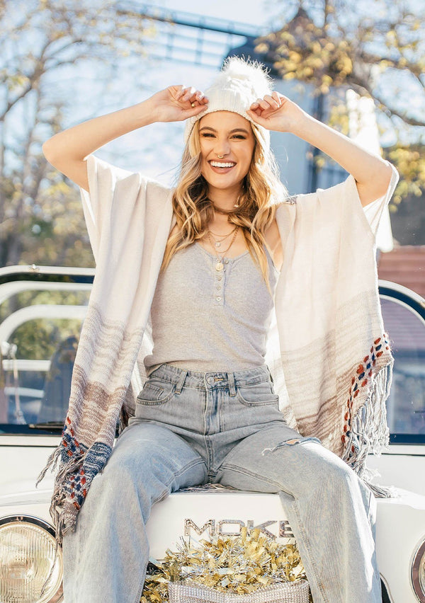 [Color: Sand/Grey] A blond woman wearing a lightweight hooded poncho. Featuring a fringed asymmetric hemline, an oversize hood, and a multi color striped pattern.