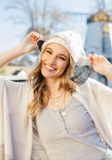 [Color: Sand/Grey] A blond woman wearing a lightweight hooded poncho. Featuring a fringed asymmetric hemline, an oversize hood, and a multi color striped pattern.