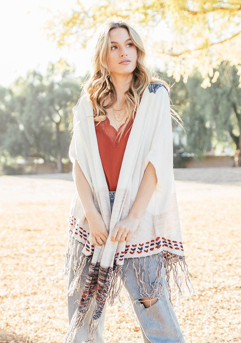 [Color: Off White/Beige] A blond woman wearing a lightweight hooded poncho. Featuring a fringed asymmetric hemline, an oversize hood, and a multi color striped pattern.