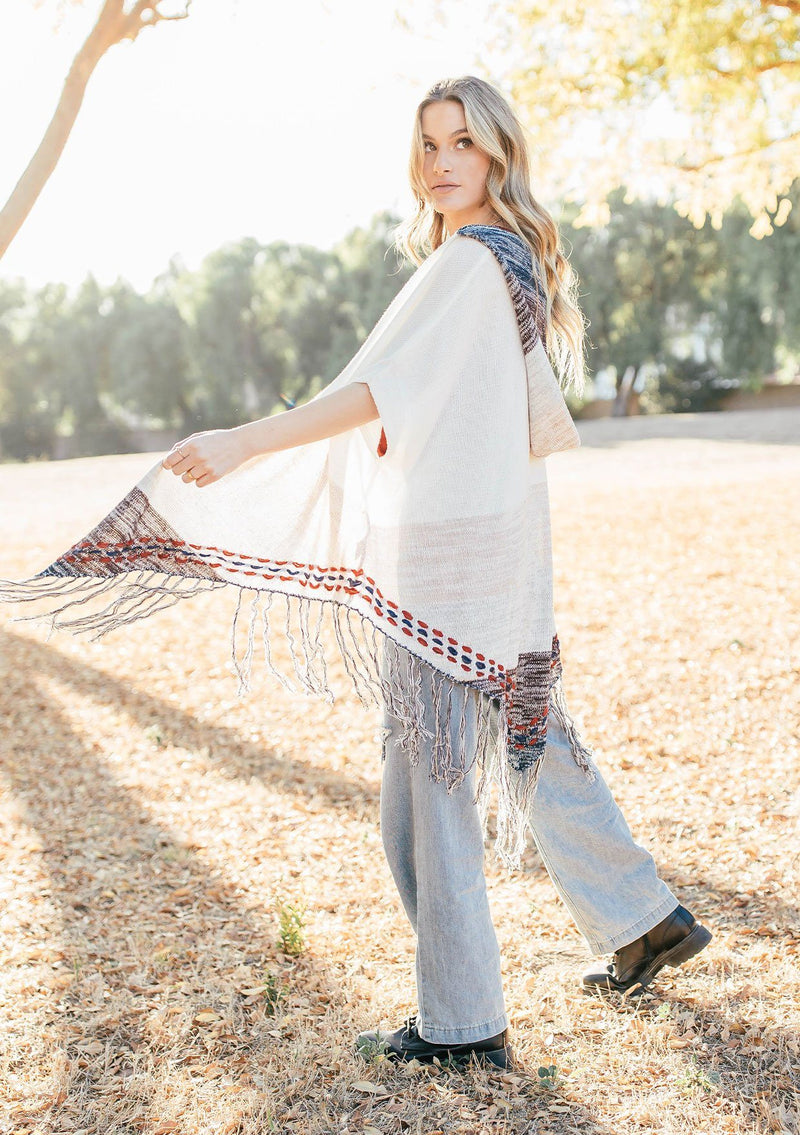 [Color: Off White/Beige] A blond woman wearing a lightweight hooded poncho. Featuring a fringed asymmetric hemline, an oversize hood, and a multi color striped pattern.