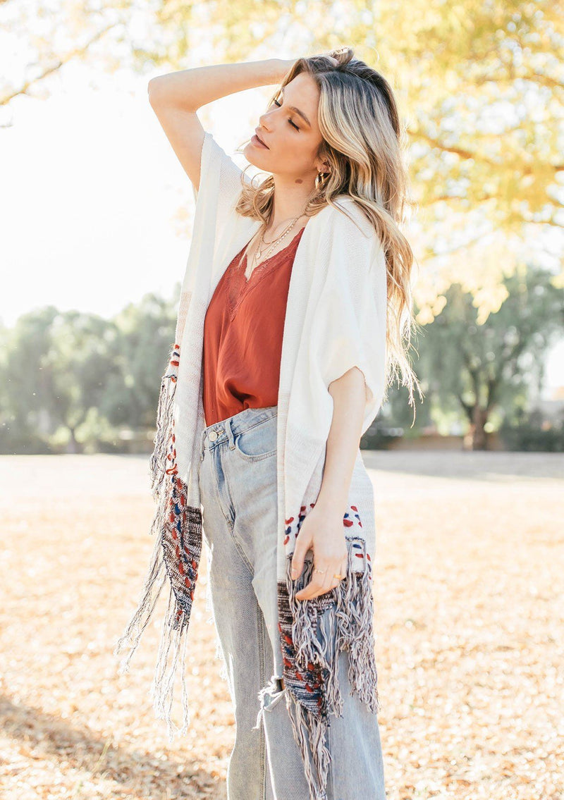 [Color: Off White/Beige] A blond woman wearing a lightweight hooded poncho. Featuring a fringed asymmetric hemline, an oversize hood, and a multi color striped pattern.