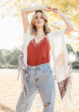 [Color: Off White/Beige] A blond woman wearing a lightweight hooded poncho. Featuring a fringed asymmetric hemline, an oversize hood, and a multi color striped pattern.