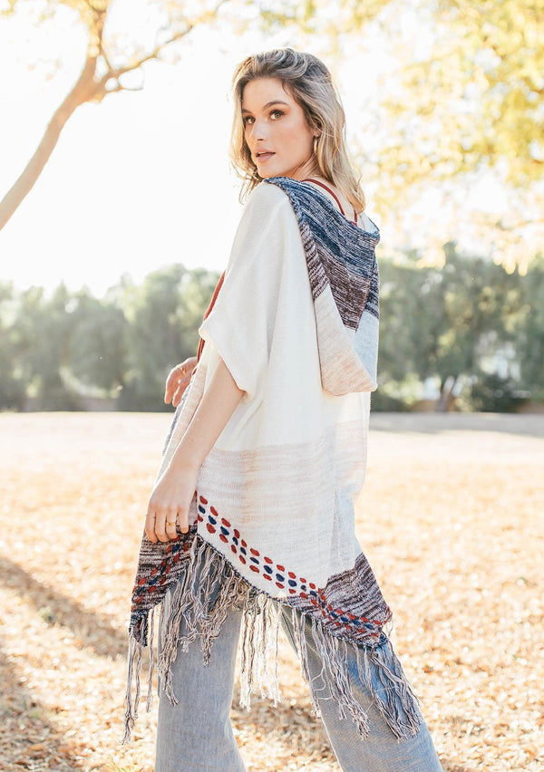 [Color: Off White/Beige] A blond woman wearing a lightweight hooded poncho. Featuring a fringed asymmetric hemline, an oversize hood, and a multi color striped pattern.