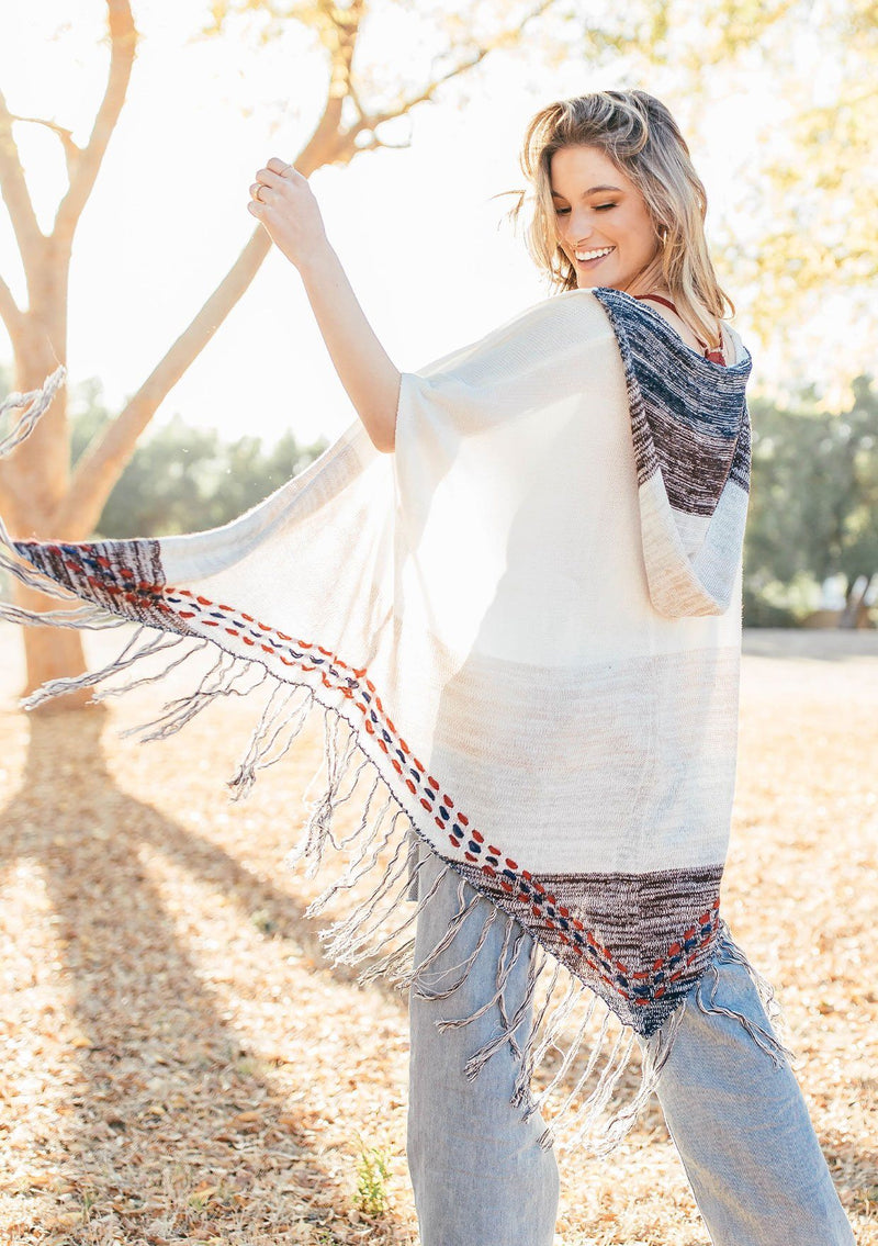 [Color: Off White/Beige] A blond woman wearing a lightweight hooded poncho. Featuring a fringed asymmetric hemline, an oversize hood, and a multi color striped pattern.