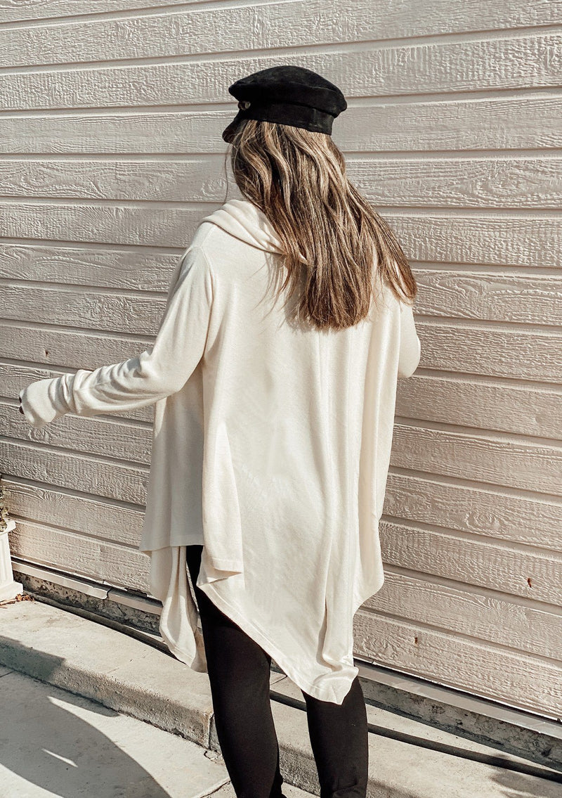 [Color: Vanilla] A woman outside wearing a cashmere blend oversize poncho sweater. Featuring a sexy deep cowl neckline, a mid length handkerchief hemline, and long sleeves with thumbholes. 