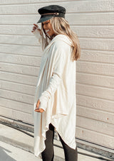 [Color: Vanilla] A woman outside wearing a cashmere blend oversize poncho sweater. Featuring a sexy deep cowl neckline, a mid length handkerchief hemline, and long sleeves with thumbholes. 