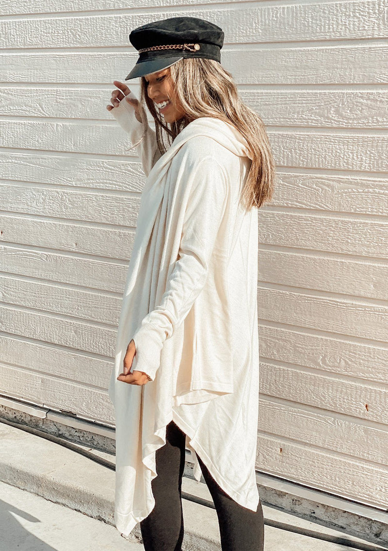 [Color: Vanilla] A woman outside wearing a cashmere blend oversize poncho sweater. Featuring a sexy deep cowl neckline, a mid length handkerchief hemline, and long sleeves with thumbholes. 