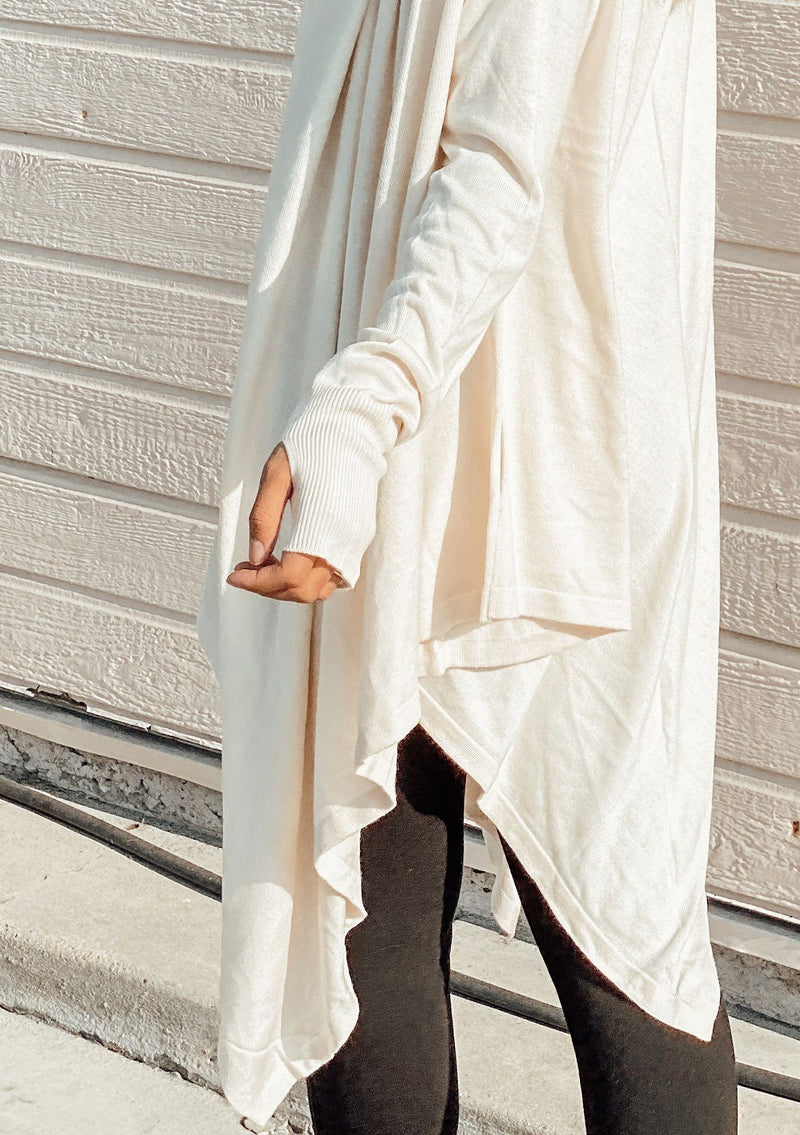 [Color: Vanilla] A woman outside wearing a cashmere blend oversize poncho sweater. Featuring a sexy deep cowl neckline, a mid length handkerchief hemline, and long sleeves with thumbholes. 