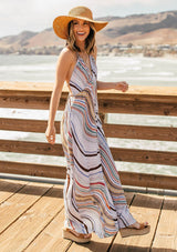 [Color: Earth Combo] A model wearing a multi color halter maxi dress in a wavy stripe print. With an open back and double strap tie, plunging v neckline, and side pockets. 