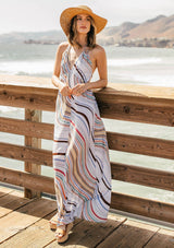 [Color: Earth Combo] A model wearing a multi color halter maxi dress in a wavy stripe print. With an open back and double strap tie, plunging v neckline, and side pockets. 