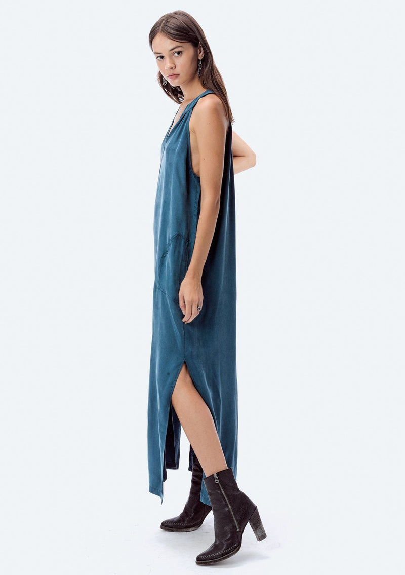 [Color: Teal] A brunette model wearing a teal cupro maxi dress with a split v-neckline, side pockets, and side slits. 