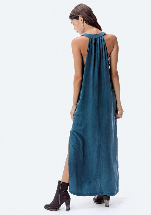 [Color: Teal] A brunette model wearing a teal cupro maxi dress with a split v-neckline, side pockets, and side slits. 