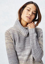 [Color: Grey/Cream] A detail image of a brunette model wearing a space dye long sleeve sweater with a slouchy turtle neck, ribbed high low hem, and side vents. A cozy fall knit sweater for casual outings and cooler temperatures. 