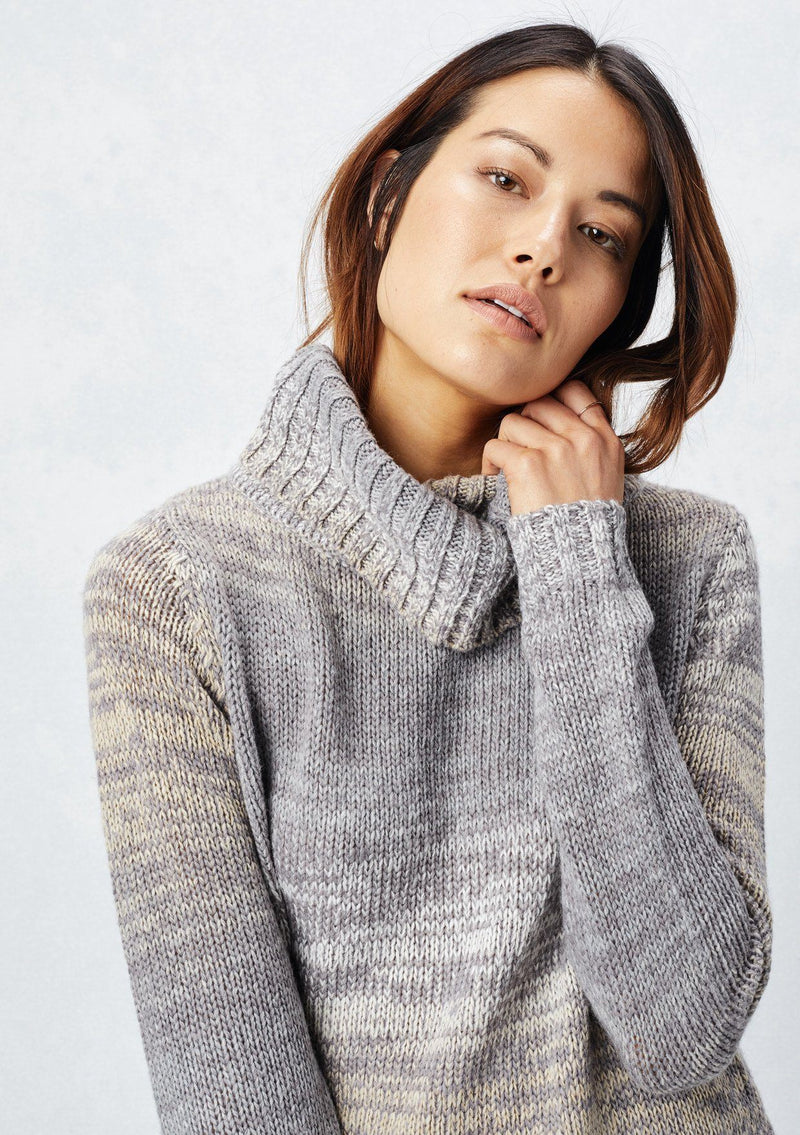 [Color: Grey/Cream] A detail image of a brunette model wearing a space dye long sleeve sweater with a slouchy turtle neck, ribbed high low hem, and side vents. A cozy fall knit sweater for casual outings and cooler temperatures. 