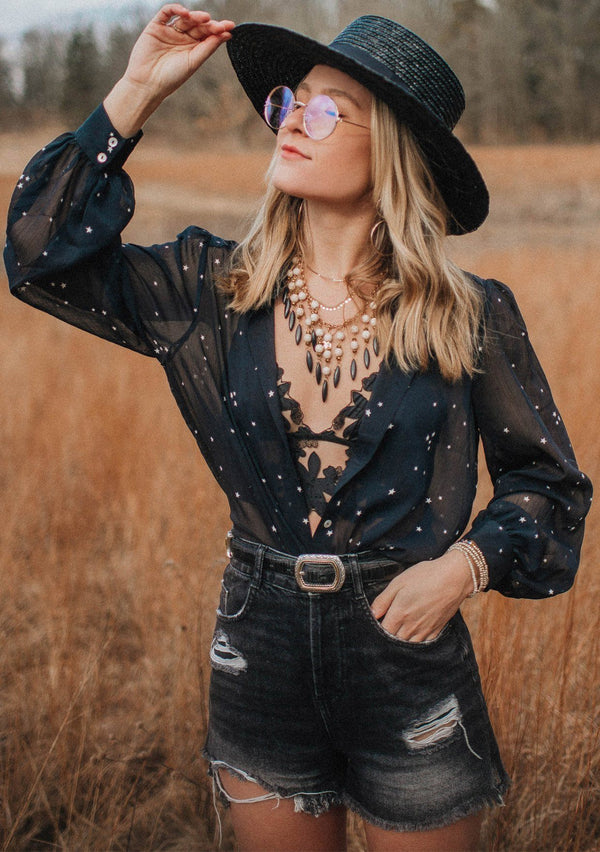 [Color: Midnight/Ivory] A blond woman wearing a sheer star printed button up shirt with volume sleeves and shirring detail, sitting with disco balls.