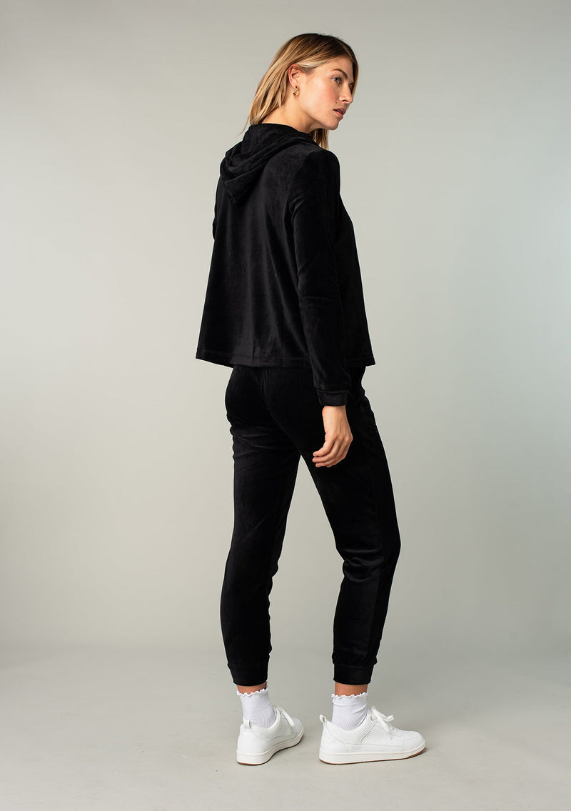 [Color: Black] A model wearing a black velour jogger pant. With a long tapered leg, side pockets, and an elastic drawstring waist. A cool retro inspired velour tracksuit.