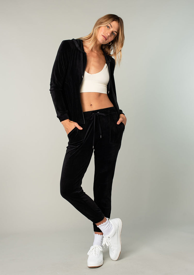 [Color: Black] A model wearing a black velour jogger pant. With a long tapered leg, side pockets, and an elastic drawstring waist. A cool retro inspired velour tracksuit.