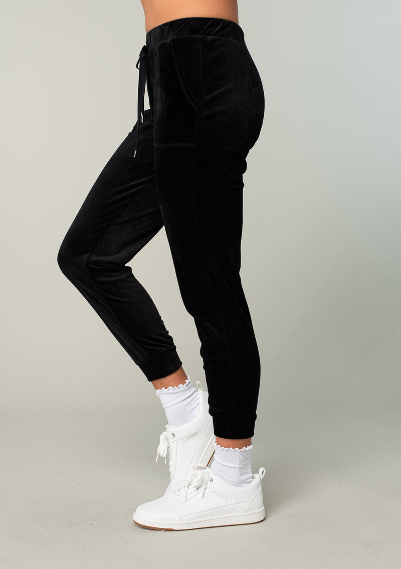 [Color: Black] A model wearing a black velour jogger pant. With a long tapered leg, side pockets, and an elastic drawstring waist. A cool retro inspired velour tracksuit.