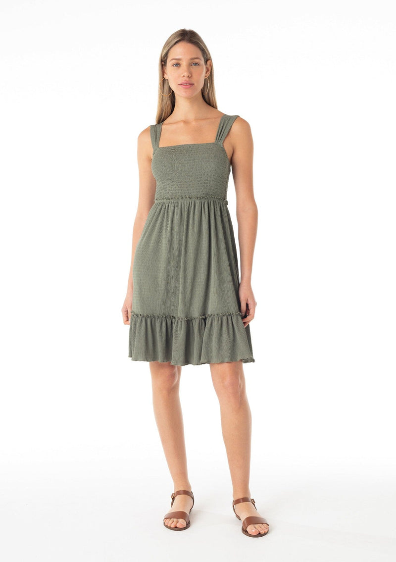 [Color: Olive] A front facing image of a blonde model wearing an olive green sleeveless spring mini dress. With wide tank top straps, a square neckline, a smocked slim fit bodice, an empire waist, and a ruffle trimmed tiered skirt.