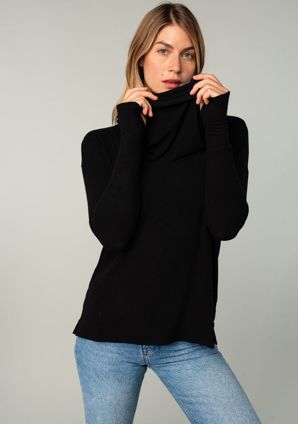 [Color: Black] A woman standing outside wearing a soft bamboo micro rib long sleeve top. Featuring an exaggerated cowl neckline that doubles as a hood, long sleeves with thumbhole accents, and breezy side vents.