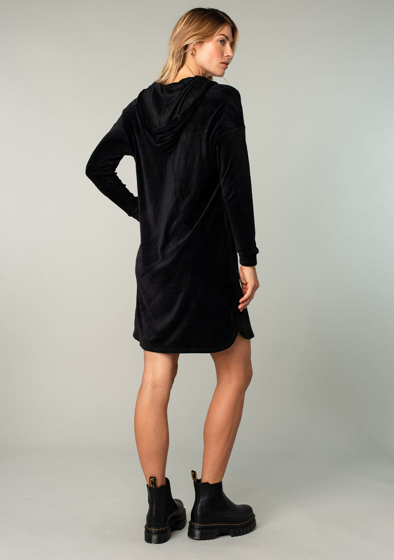 [Color: Black] A model wearing a plush black velour mini dress. With a drawstring hoodie, long sleeves, and side pockets.