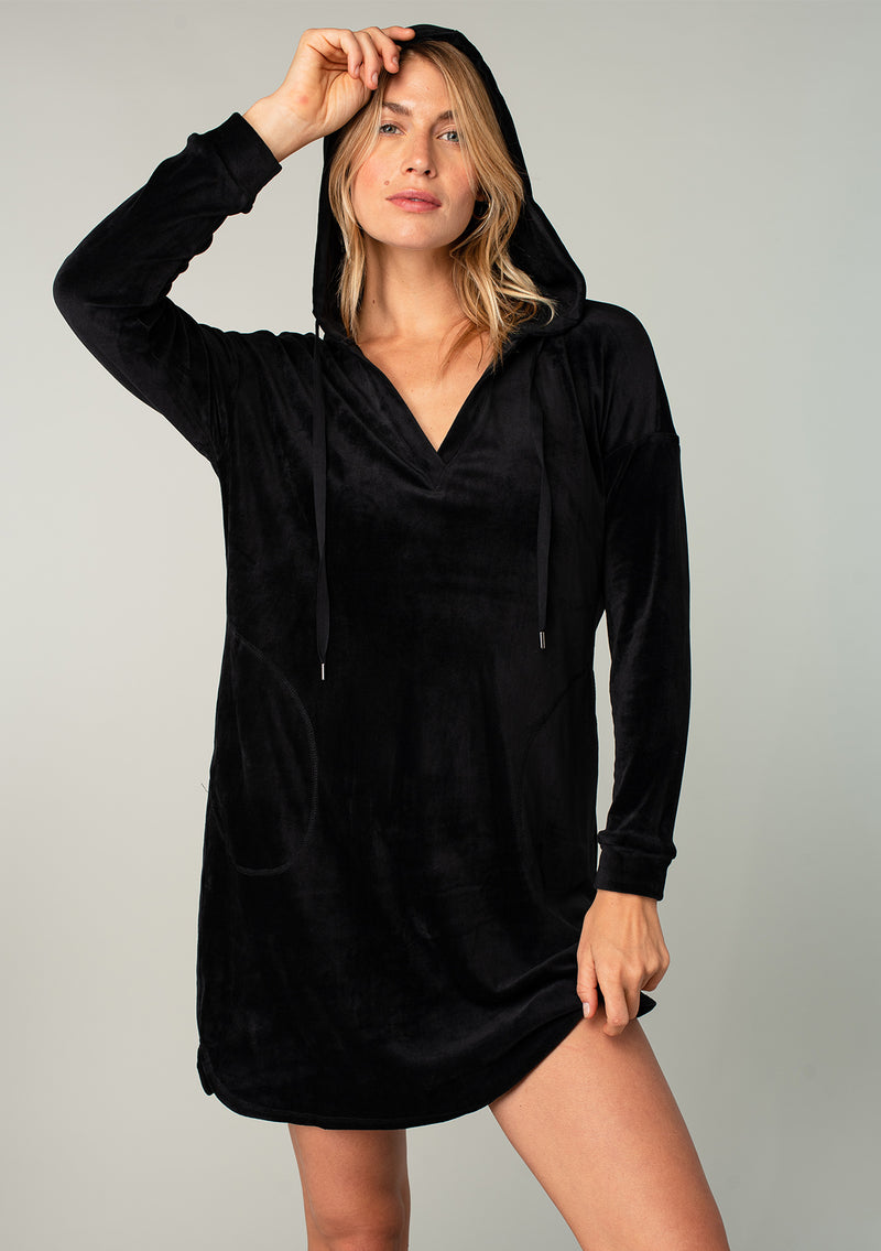 [Color: Black] A model wearing a plush black velour mini dress. With a drawstring hoodie, long sleeves, and side pockets.