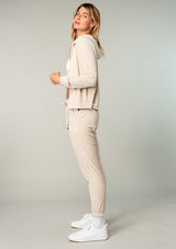 [Color: Stone] A model wearing a cream colored velour jogger pant. With a long tapered leg, side pockets, and an elastic drawstring waist. A cool retro inspired velour tracksuit.