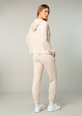 [Color: Stone] A model wearing a cream colored velour jogger pant. With a long tapered leg, side pockets, and an elastic drawstring waist. A cool retro inspired velour tracksuit.