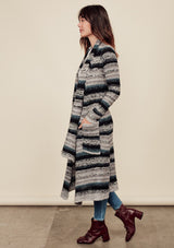 [Color: Grey/Teal/Black]  A side facing image of a brunette model wearing a bohemian striped knit duster cardigan in grey and blue with a shawl collar, side pockets, and a cozy, draped fit perfect for layering over denim.